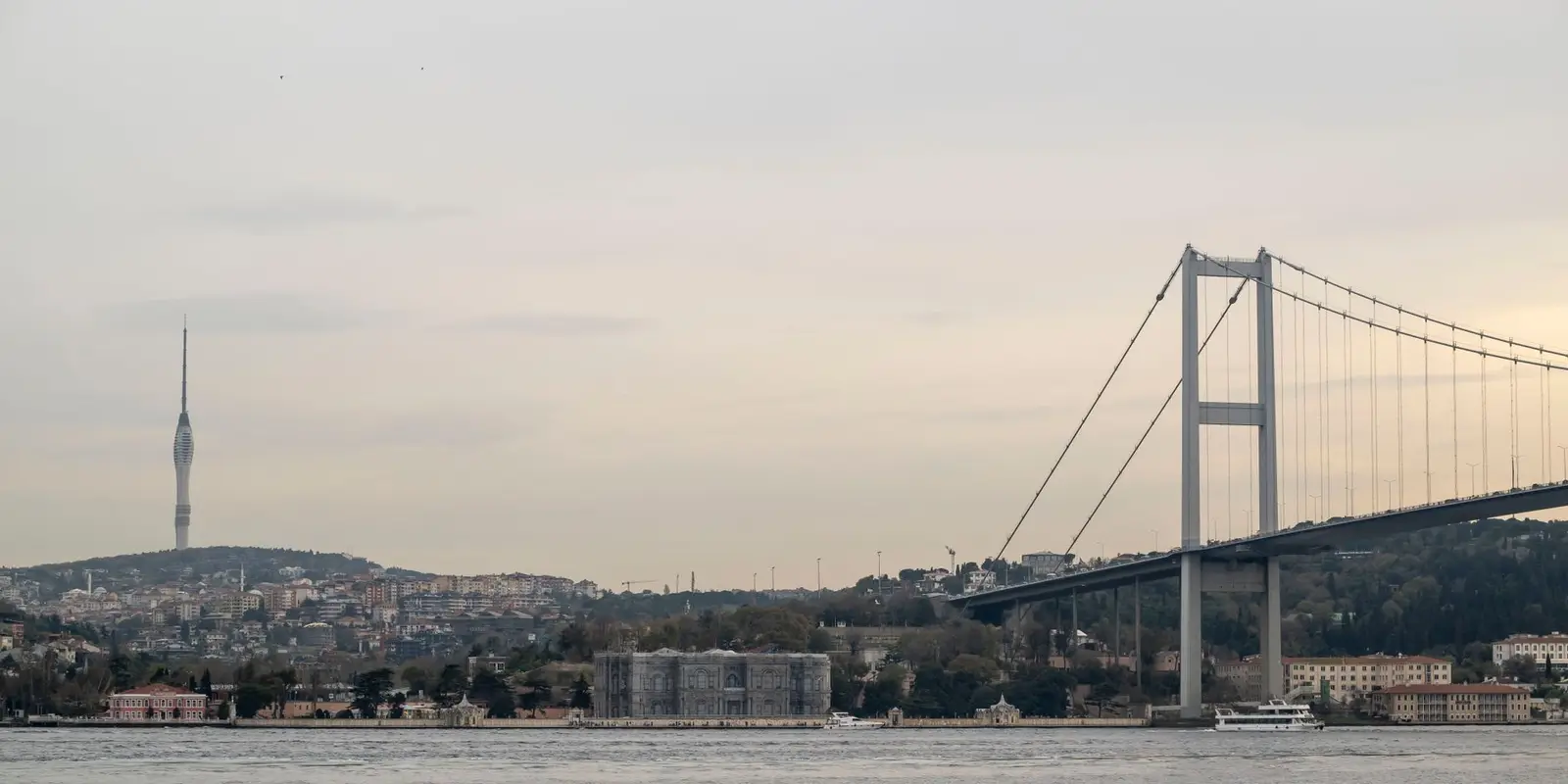 View of the Bosphorus Bridge and Istanbul skyline at dusk, featuring calm waters and cityscape.