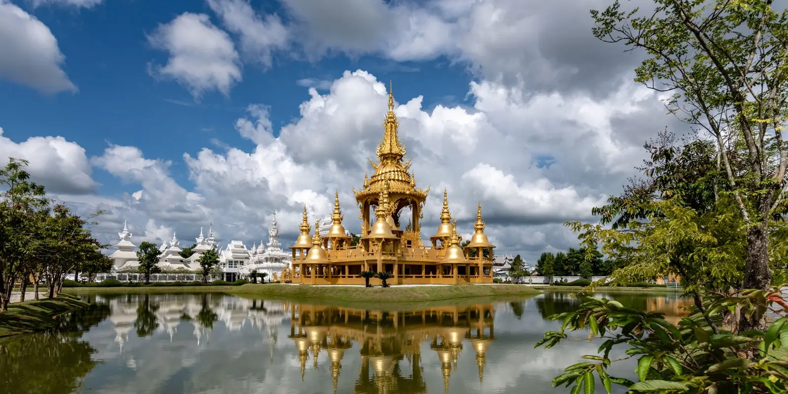 Golden Wat Rong Khun reflected in a serene lake, under a dramatic cloudy sky in Chiang Rai, Thailand.